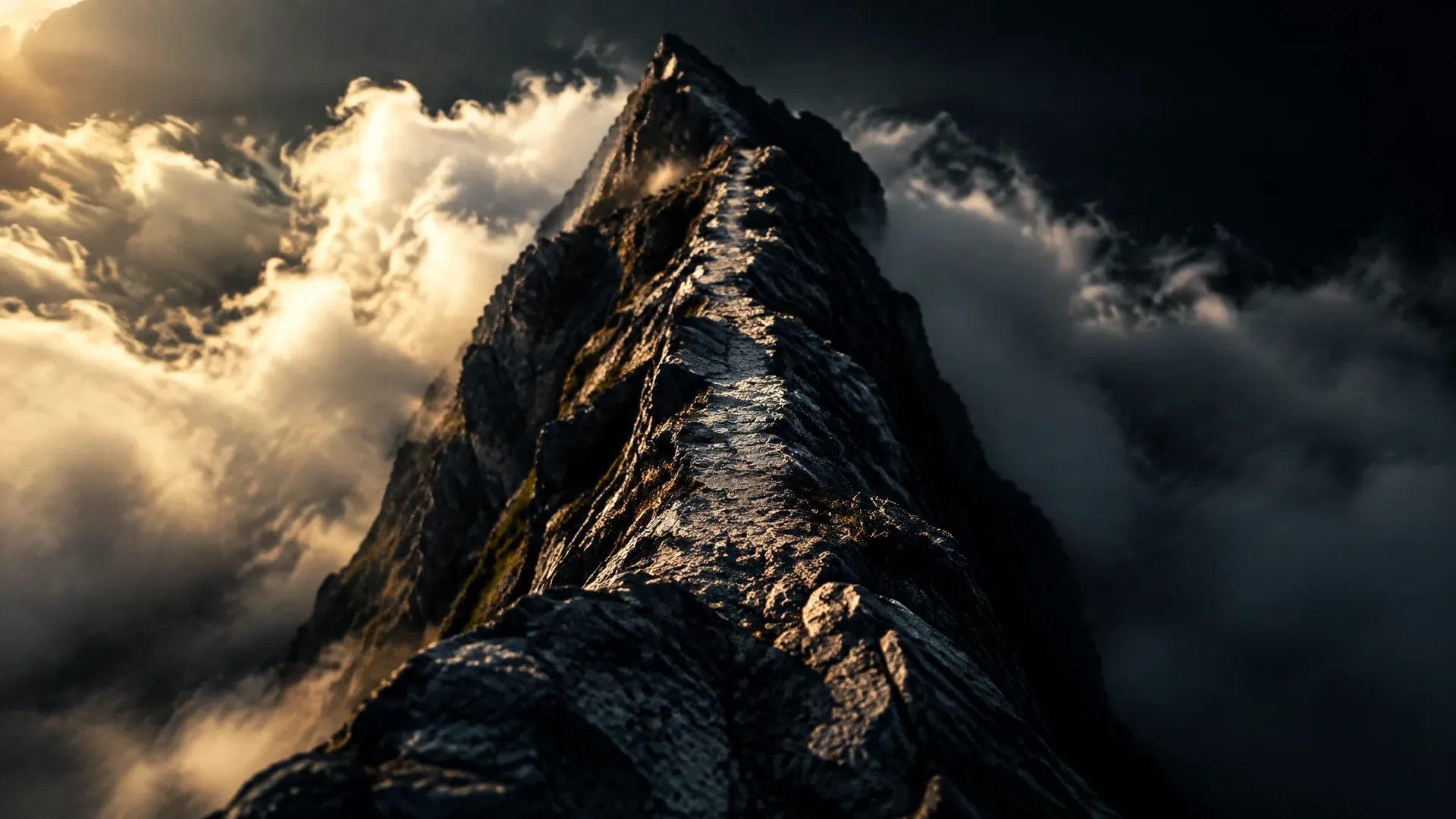 Schmaler Felsgrat aus dunklem Gestein im harten Seitenlicht, der Pfad verengt sich, die Flanken fallen beidseits in Wolken ab