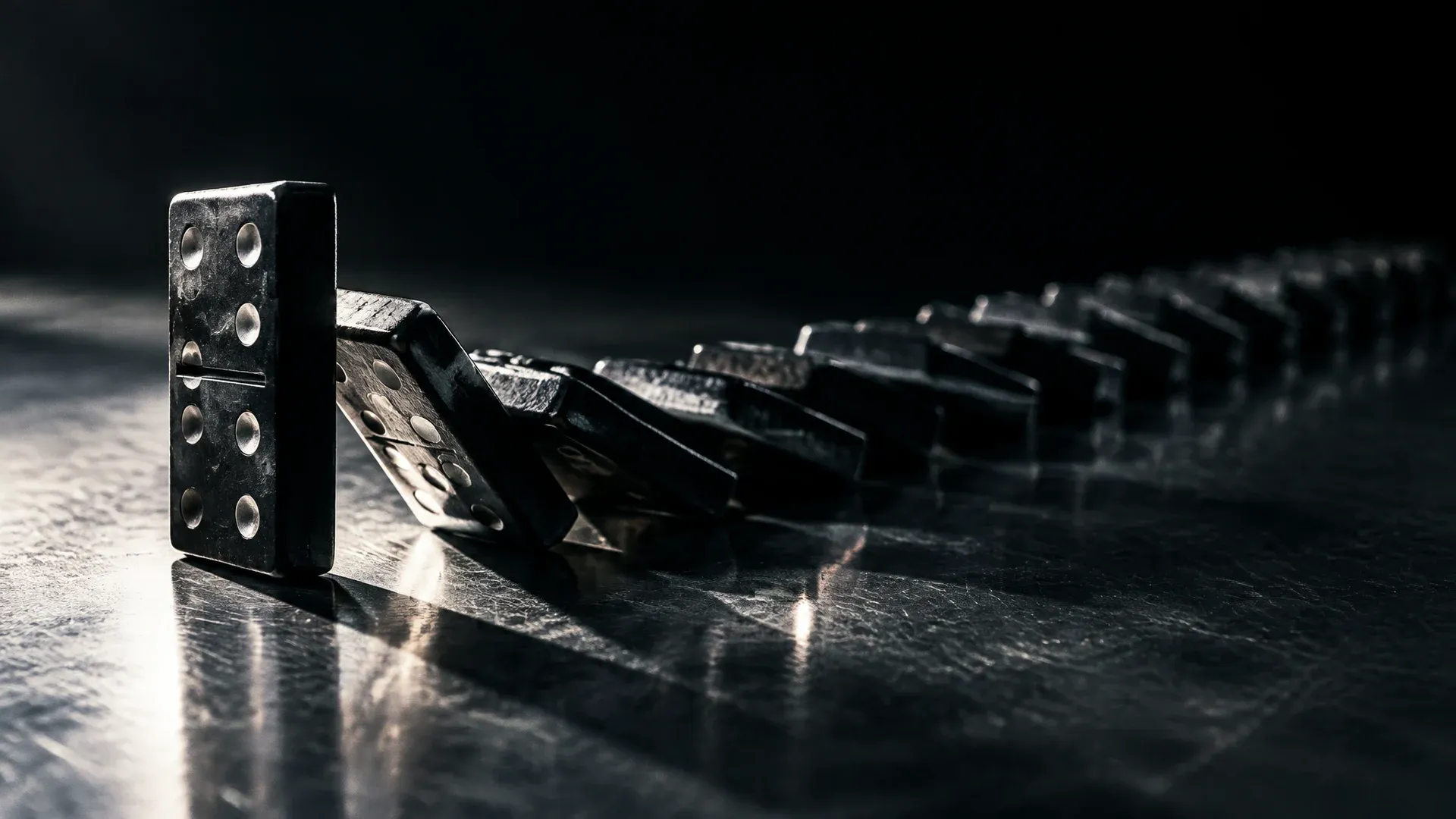 Lange Reihe dunkler Dominosteine auf einer Metallflaeche, die vorderen Steine liegen umgefallen, ein schwerer Stein am Anfang der Reihe steht noch aufrecht