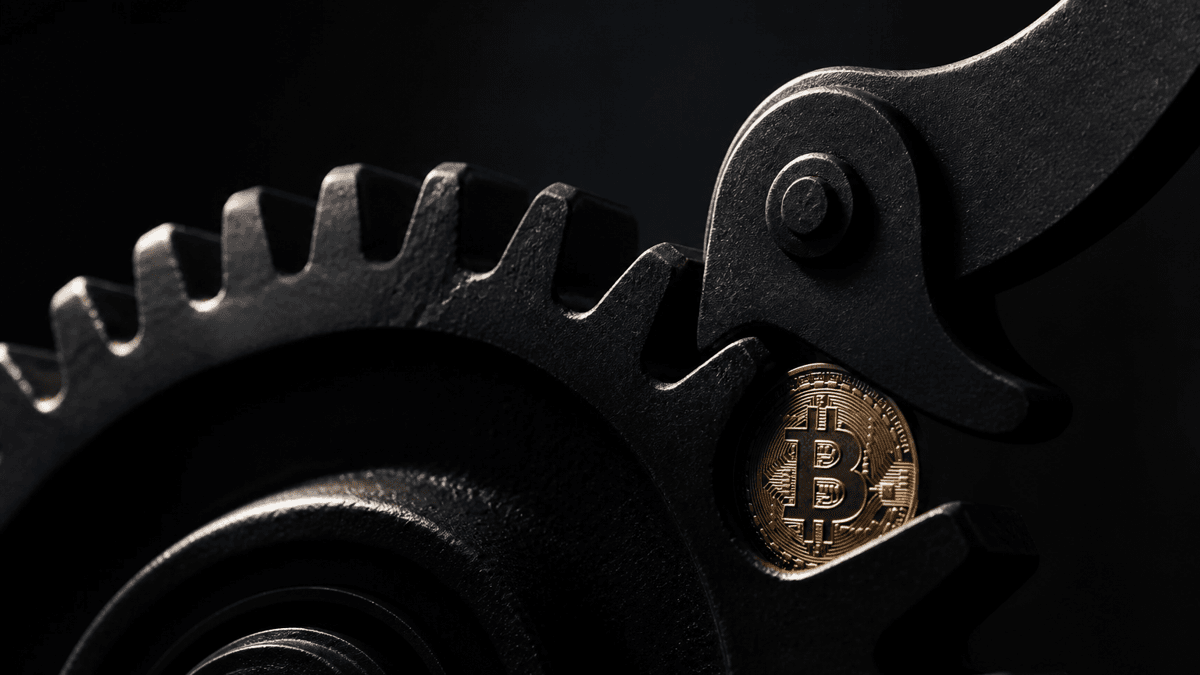 Ein Stahlzahnrad mit Sperrklinke, die nur eine Drehrichtung zulaesst, mit einer eingeklemmten Muenze mit Bitcoin-Symbol