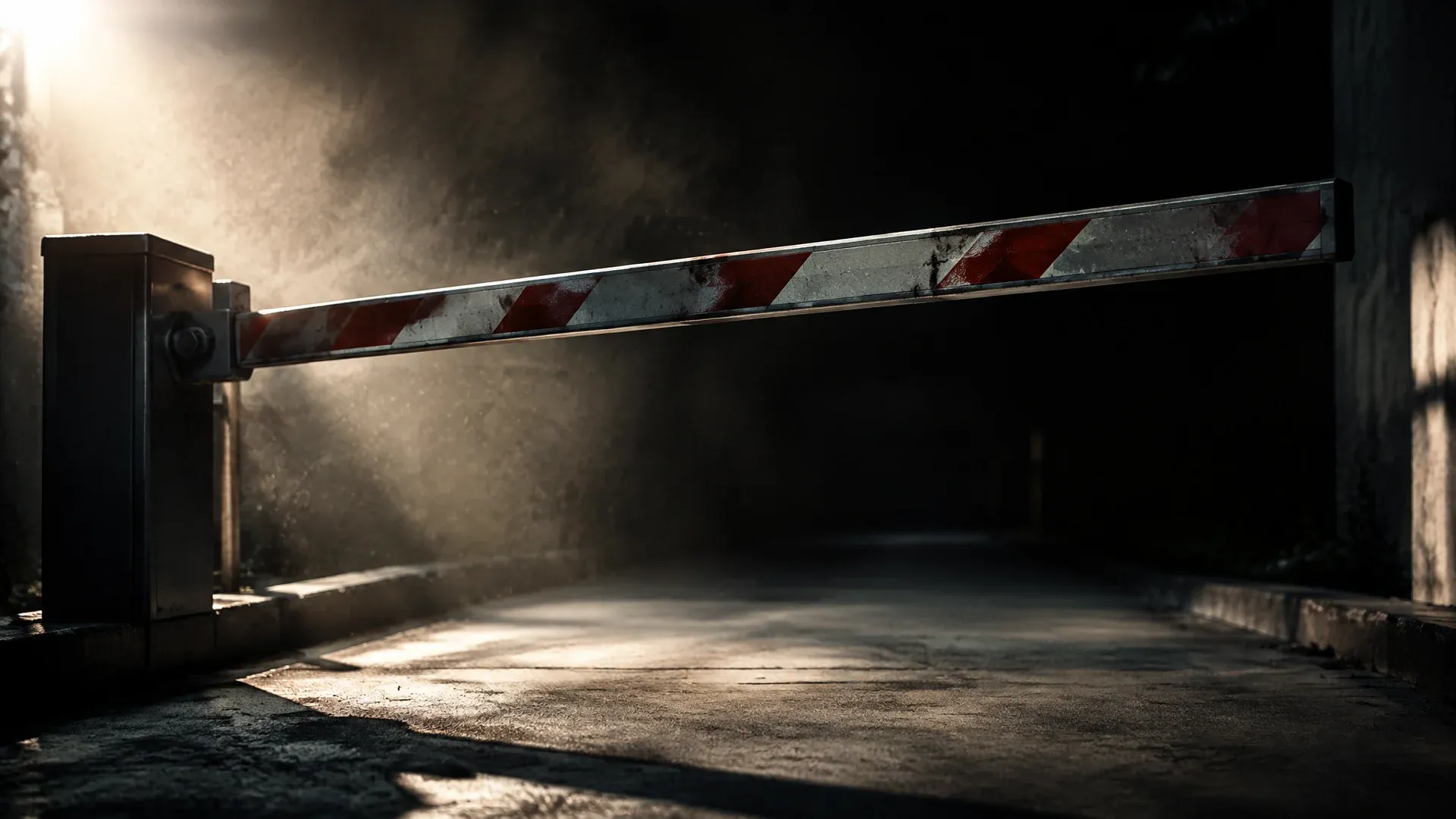 Schwere Stahlschranke senkt sich im Gegenlicht über eine leere Betonzufahrt, feiner Staub steht in der Luft