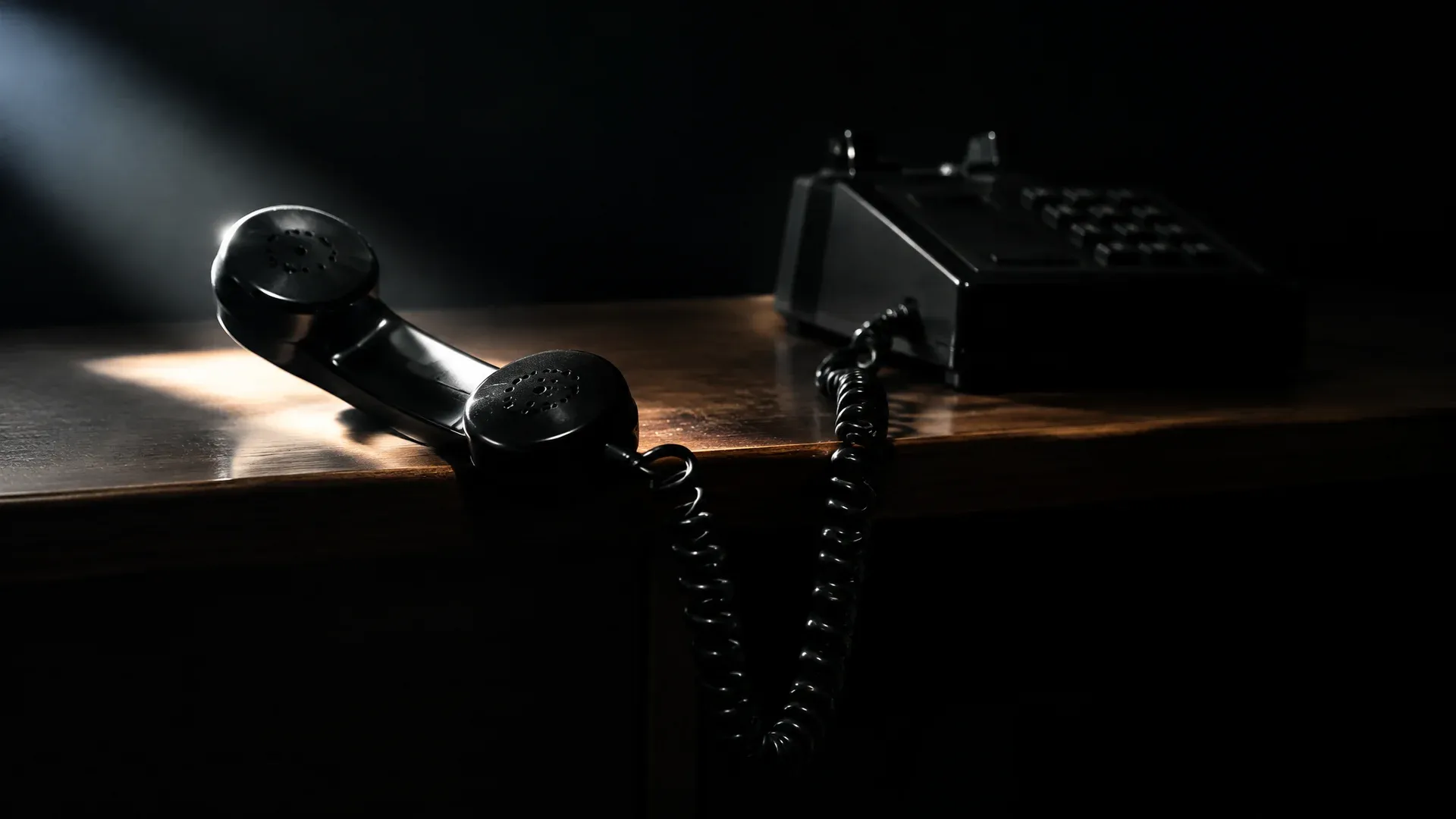 Schwarzes Tischtelefon mit abgenommenem Hörer auf einem leeren Schreibtisch im Halbdunkel