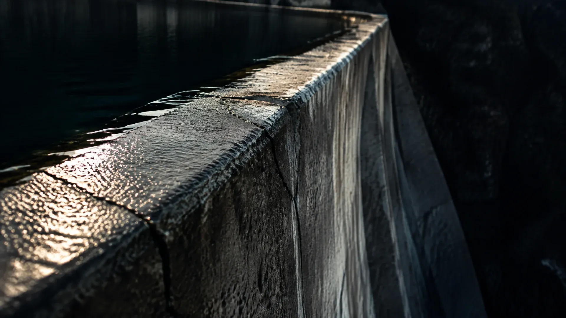 Betonkrone eines Stauwehrs im Streiflicht, das dunkle Wasser steht wenige Zentimeter unter der Kante und liegt still