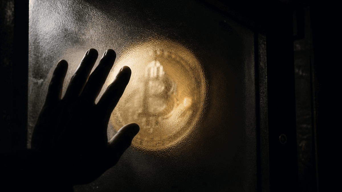 Coin stamped with a Bitcoin symbol behind a frosted glass pane, a shadowy hand in front of it