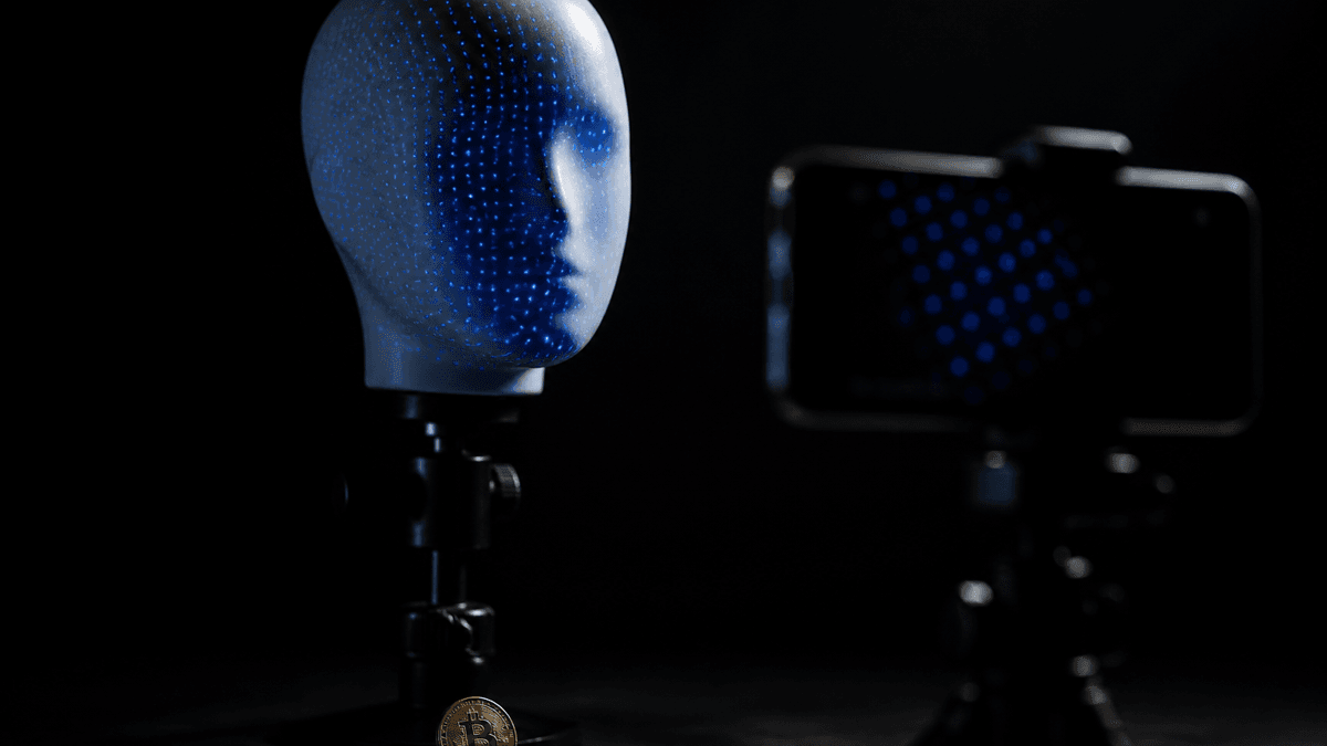 Ein gesichtsloser Puppenkopf wird von einem Punktraster aus blauem Licht abgetastet, davor ein Smartphone auf einem Stativ und eine Münze mit Bitcoin-Zeichen