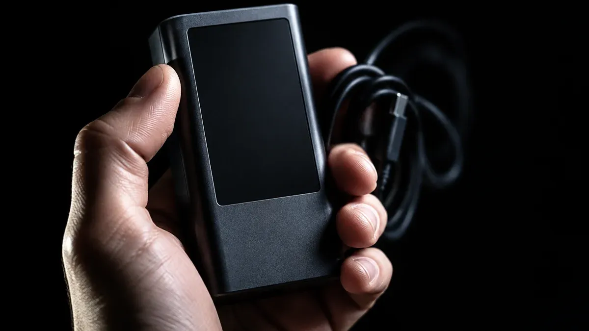 Hand hält ein schmuckloses dunkelgraues Elektronikgerät mit erloschenem Display und aufgerolltem Kabel