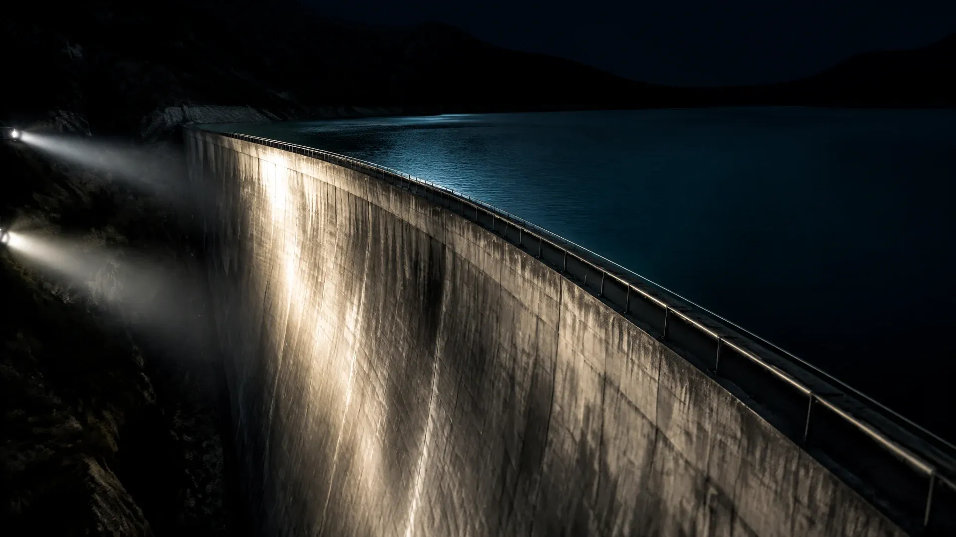 Betonstaumauer eines randvollen Speicherbeckens bei Nacht, von der Seite angestrahlt