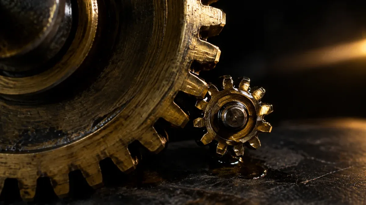 Macro shot of two interlocking brass gears, one very large and a tiny one beside it, on oily gleaming workshop steel.