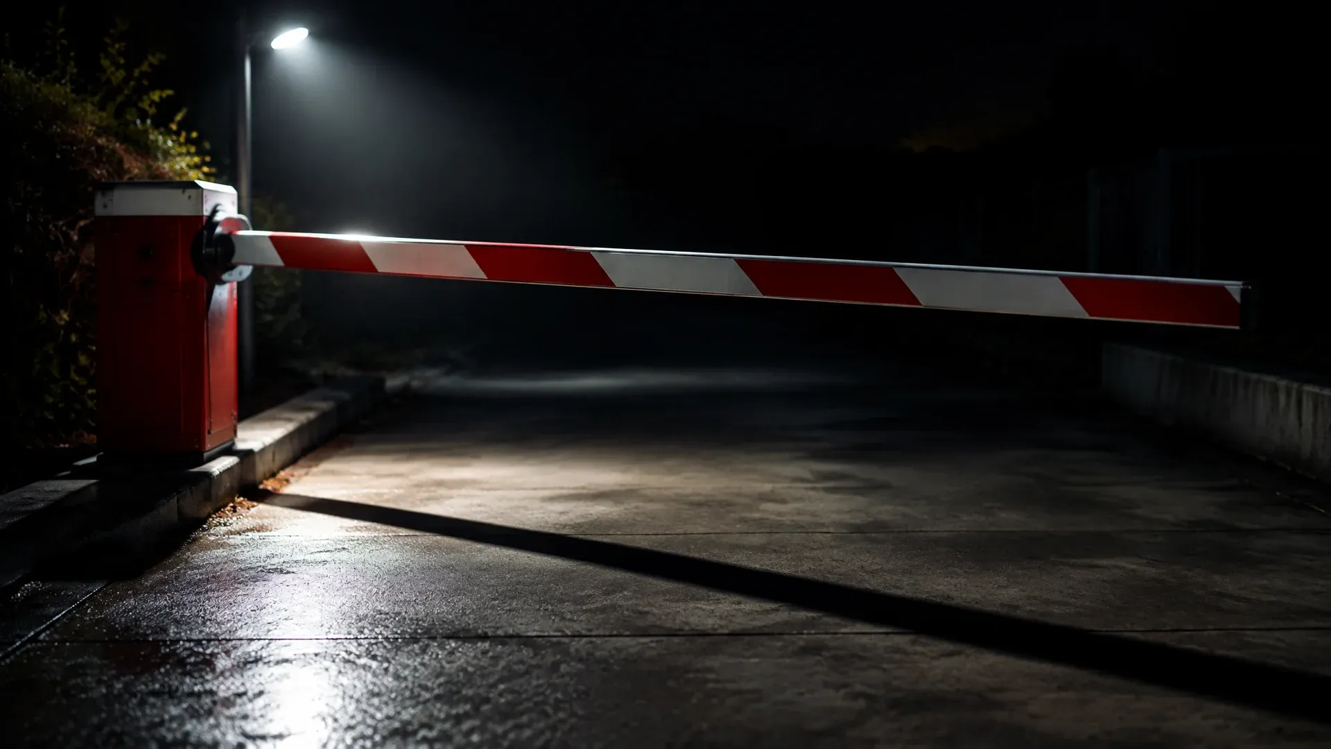 Rot-weiß lackierte Schranke senkt sich über eine leere nächtliche Betonzufahrt, ein Scheinwerfer wirft den langen Schatten des Schrankenbaums