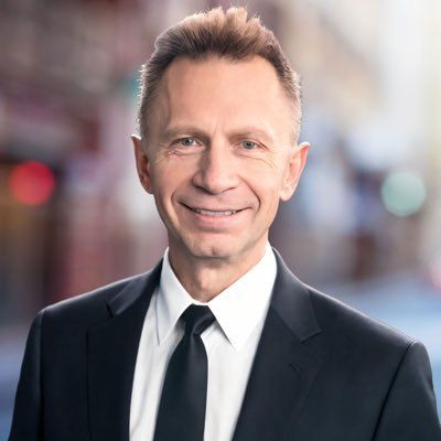 Portrait of Wojciech Kaszycki in a dark suit with a white shirt and black tie in front of a blurred street scene