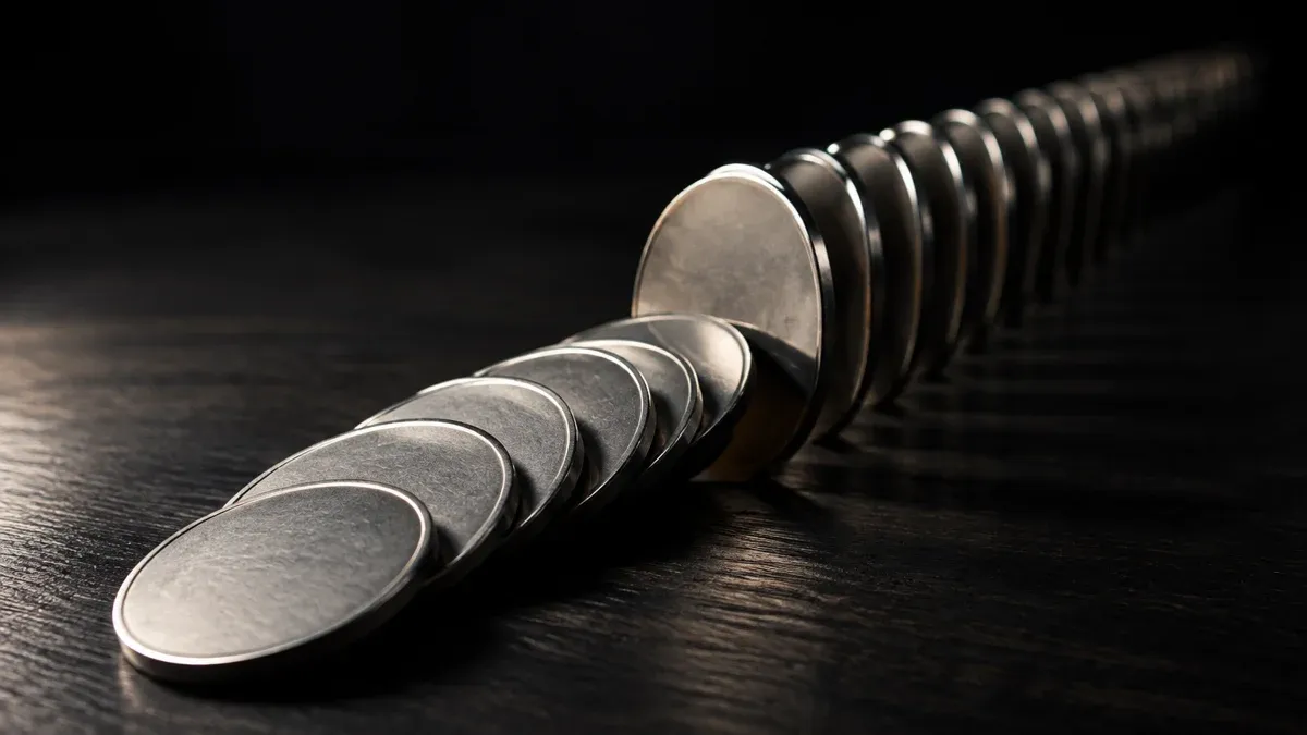 A long row of metal coins standing on edge on a dark wooden surface, the front seven fallen over and lying on top of each other